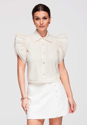 Nainen pukeutuneena valkoiseen puseroon, jossa on suuret röyhelöiset hihat, ja valkoiseen minihameeseen, jossa on sivulla napit. Hänellä on kultaiset korvakorut ja rannekorut asusteina.