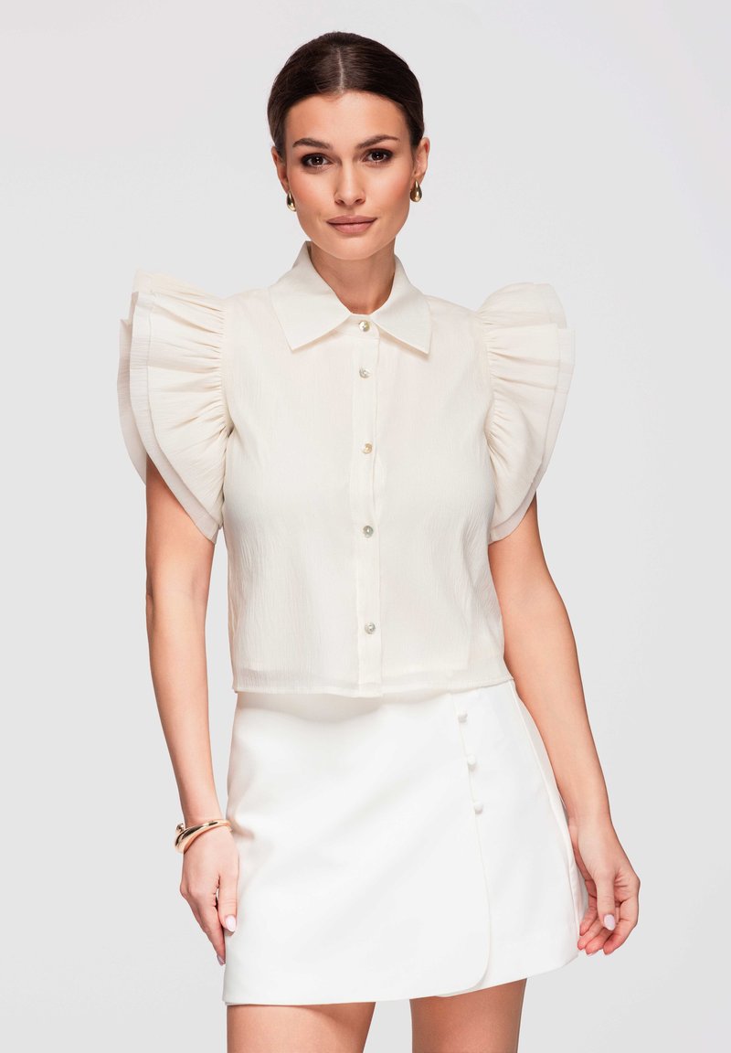 Femme portant un chemisier blanc à larges manches volantées et une mini-jupe blanche à boutons latéraux, accessoirisée avec des boucles d'oreilles et des bracelets dorés.