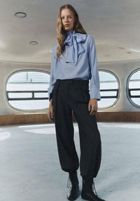 Blusa azzurra chiara con un grande fiocco al colletto, abbinata a pantaloni scuri a vita alta e scarpe alla caviglia nere con lacci.