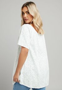 White short-sleeve top with a textured floral pattern, round neckline, and a relaxed fit, paired with blue denim jeans.