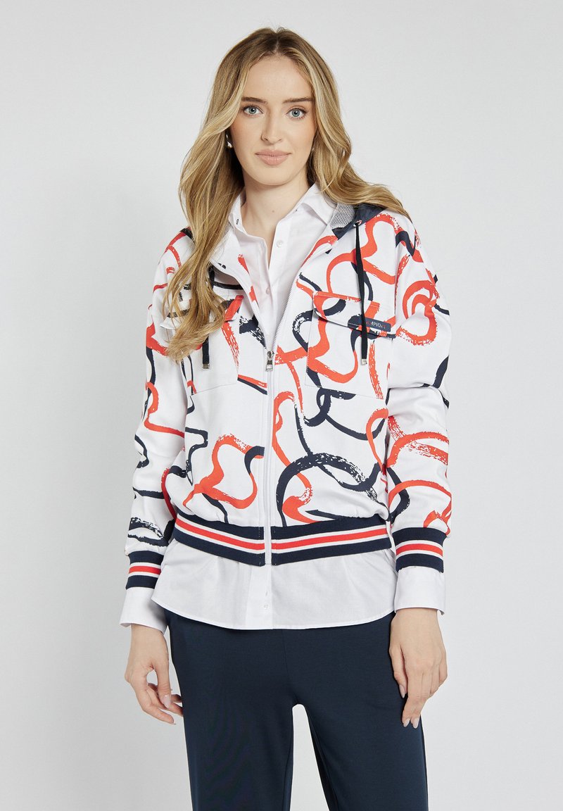 Biała bluza z zamkiem błyskawicznym z czerwonym i granatowym abstrakcyjnym wzorem, dwie przednie kieszenie oraz granatowe paski na mankietach i u dołu. Nosi się ją na białej koszuli.