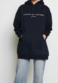 Sudadera con capucha de color azul marino con el logo blanco "TOMMY HILFIGER", bolsillo frontal y cordones. La tela es suave y lisa, con un corte casual.