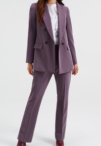 Lila dubbelknäppt kavaj med svarta knappar, i kombination med matchande skräddarsydda byxor, och en vit skjorta som syns under. Slät tygtextur.