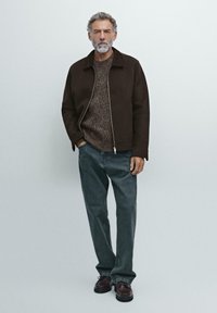 Giacca marrone con zip sopra un maglione di maglia marrone, jeans grigi e scarpe scure. La giacca presenta un colletto strutturato e una texture liscia.