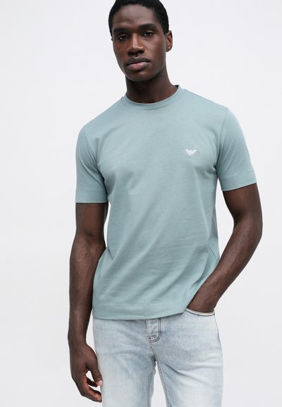 Svijetlozeleni T-shirt kratkih rukava od mekog pamučnog materijala s okruglim izrezom i malim logom na lijevoj strani prsnog koša. Uparen s svijetloplavim trapericama.