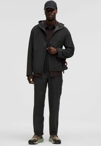 Čierna nepremokavá bunda s kapucňou, skombinovaná s čiernym tričkom a tmavými nohavicami. Osoba má na hlave námornícku čiapku a svetlohnedé tenisky.