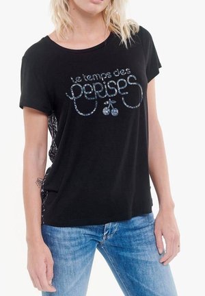Vrouw draagt een zwart T-shirt met Franse tekst 'le temps des cerises' en een afbeelding van kersen, gecombineerd met lichtblauwe jeans tegen een witte achtergrond.