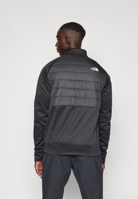 Chaqueta negra con una sección acolchada en la parte superior de la espalda, una sección inferior de tela suave y un logo en el hombro.