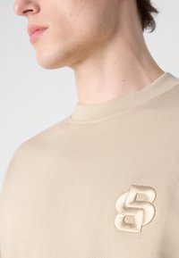 Beige textúrájú pulóver bordás mintával, világos színű hímzett logóval a mellkason és kerek nyakkal.