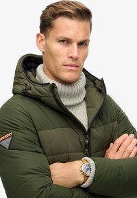 Chaqueta tipo puffer en verde oliva con capucha y cremallera negra. Lleva un cuello alto de punto gris debajo y un reloj con correa de cuero marrón.