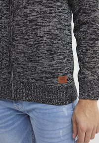 Cárdigan de punto negro y gris con cremallera, patrón texturizado y una etiqueta de cuero marrón en la parte inferior derecha. Llevado sobre unos jeans azules.