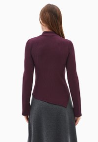 Długi rękaw, dopasowana bluzka w głębokim purpurowym ryflowanym materiale, z wysokim dekoltem i asymetrycznym dołem, zestawiona z szarą spódnicą.