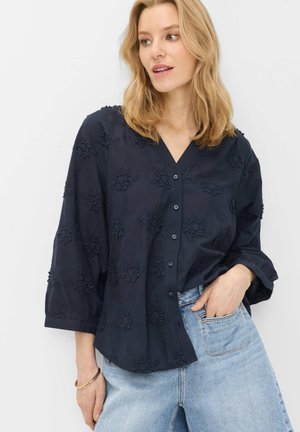 Kvinde med blondt hår har en løs mørkeblå knapbluse med blomstertekstur og lyseblå jeans på, poserer med den ene hånd i lommen.