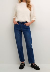 Maglione bianco a maglia con maniche corte, abbinato a jeans di jeans blu a vita alta e scarpe nere con dettaglio di catena dorata.