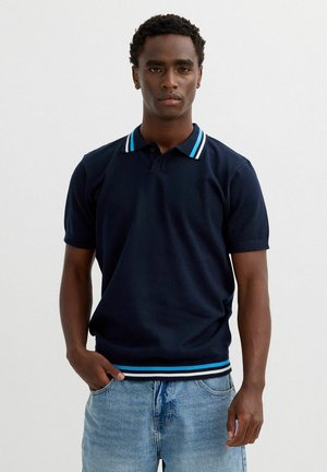 Joven de pie con una mano en el bolsillo, lleva una camiseta polo azul marino con cuello y dobladillo a rayas, combinada con vaqueros azul claro.