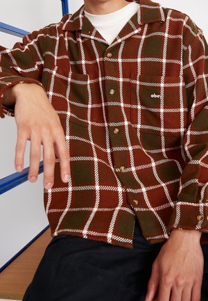 Man draagt een bruin en olijfgroen geruit overhemd met knopen en een wit onderhemd, zittend met één arm uitgestrekt en een ontspannen houding.