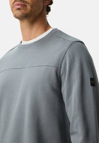 Grijze trui van gladde stof, met een ronde halslijn, een horizontale naad over de borst en een logopatch op de mouw.