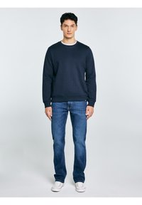 Młody mężczyzna ubrany w granatową bluzę, niebieskie dżinsy i białe trampki, stojący na tle jednolitego, jasnoszarego tła.