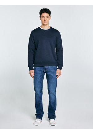 Jauns vīrietis tumsā zils sporta krekls, zili džinsi un balti sporta apavi stāv pret vienkrāsainu gaiši pelēku fonu.