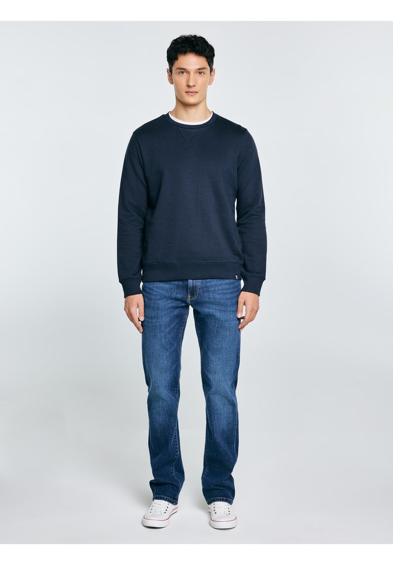 Młody mężczyzna ubrany w granatową bluzę, niebieskie dżinsy i białe trampki, stojący na tle jednolitego, jasnoszarego tła.