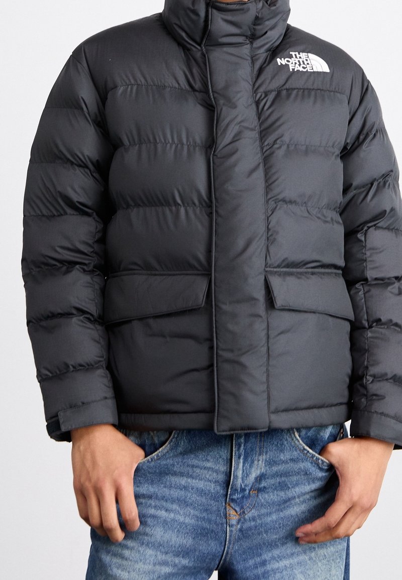 Gewatteerde zwarte jas met een hoge kraag, uitgerust met zijzakken en een sluiting aan de voorkant. Het North Face-logo is zichtbaar aan de bovenkant van de linkerzijde.