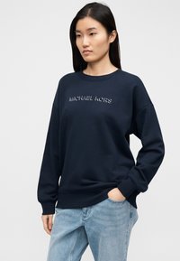 Tamsiai mėlynas džemperis su apvalia iškirpte, puoštas sidabriniu siuvinėjimu „MICHAEL KORS“. Ribinės rankogaliai ir apačia. Derinamas su šviesiai mėlynomis džinsų kelnėmis.