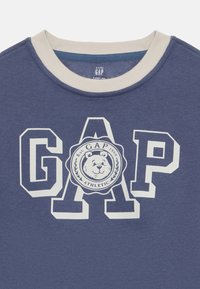 Blauer Sweatshirt mit einem runden Bärenlogo und "GAP" in großen weißen Buchstaben. Verfügt über einen hellcremefarbenen, gerippten Rundhalsausschnitt und eine weiche Stofftextur.