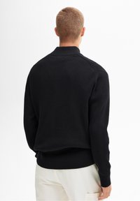 Black knit sweater featuring a ribbed texture, high collar, and fitted cuffs, worn with light-colored trousers.