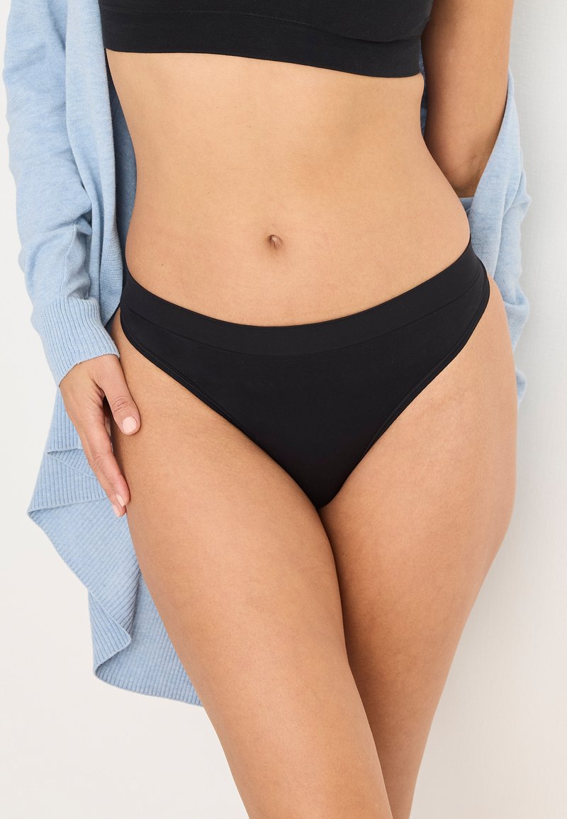 Zwarte bikini met een hoge cut en een gladde textuur, aangevuld met een zachte, lichtblauwe cardigan die aan de zijkant hangt.