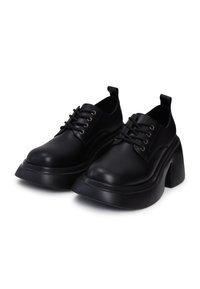 Paire de chaussures à plateforme épaisses noires à lacets, avec bouts arrondis et languettes au talon sur fond blanc.