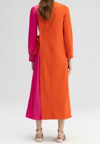Femme portant une longue robe avec un côté orange et un côté rose, debout pieds nus sur un fond blanc uni.