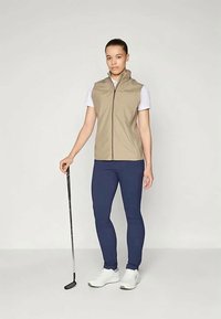 Beige bezpiedurkņu veste ar priekšējo rāvējslēdzēju, apvienota ar pieguļošām tumši zilām legingām un baltām kurpēm. Modelis tur golfu nūju. Gluda tekstūra.
