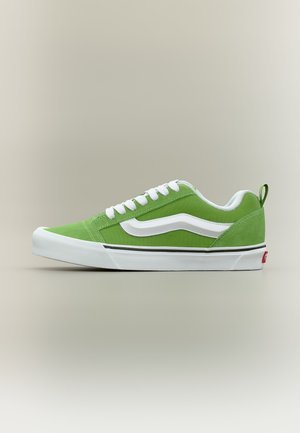 Groene lage sneaker met witte veters, witte zijstreep en witte zool met zwarte streep, in profiel afgebeeld tegen een neutrale achtergrond.