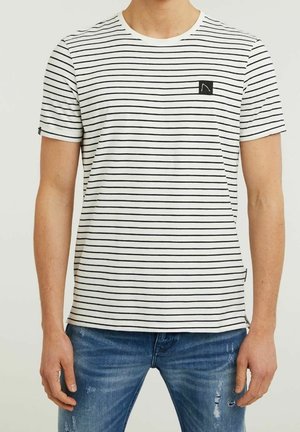 Mand iført en hvid kortærmet t-shirt med tynde sorte horisontale striber og blå slidte jeans, stående med arme afslappede.