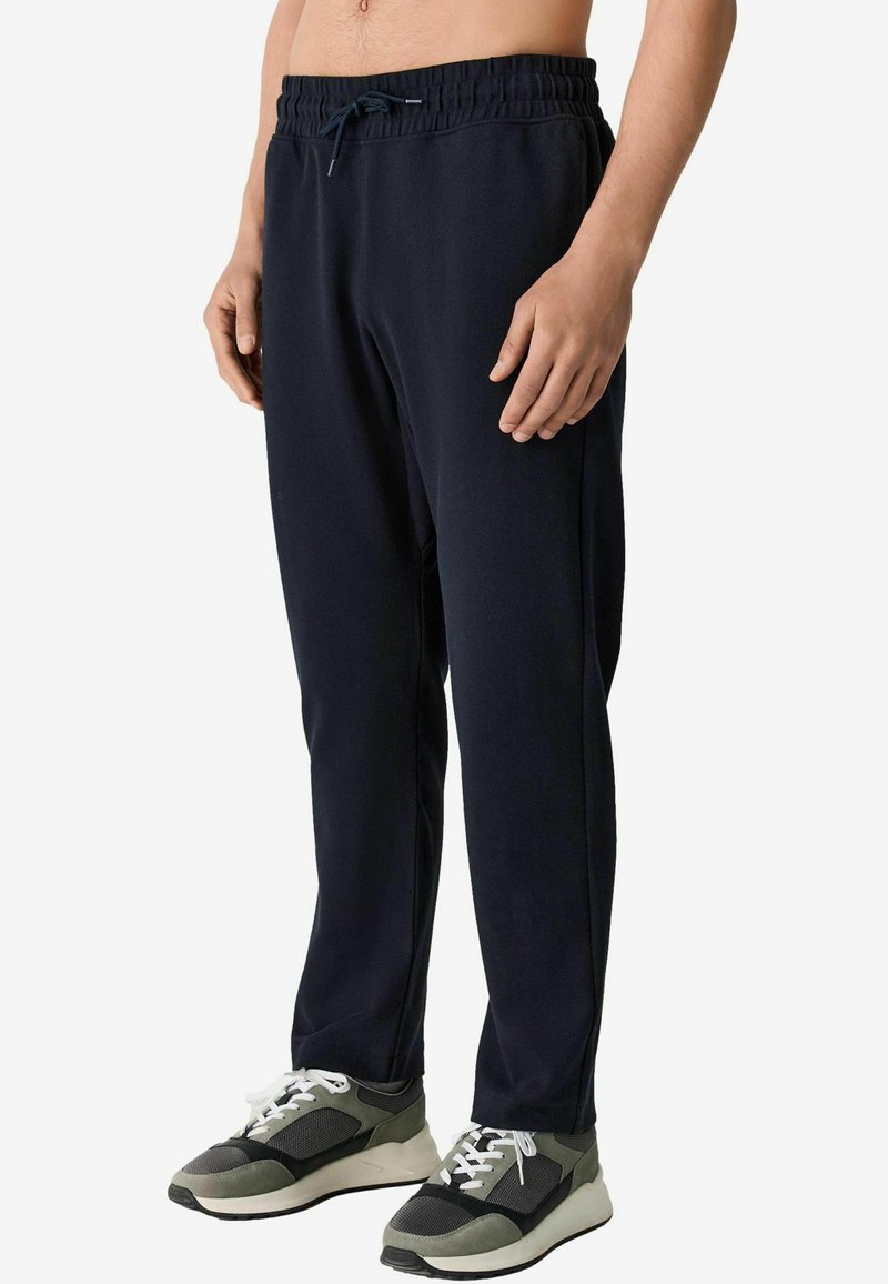 Mann trägt schwarze Sweatpants mit Kordelzug und graue, weiße und schwarze Sportschuhe, steht vor weißem Hintergrund.