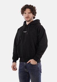 Felpa nera in cotone, con tasca frontale, cappuccio regolabile e piccolo logo bianco sul petto. Indossata con jeans blu.