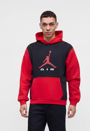 Ung mand iført en sort og rød hættetrøje med en rød basketball-logo og ordet AIR, stående mod en ensartet lys baggrund.