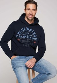 Sudadera azul marino con un diseño gráfico frontal en azul claro y texto rojo. Cuenta con capucha con cordón y bolsillo tipo canguro. Se lleva con jeans azul claro.