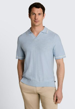 Mężczyzna w jasnoniebieskim dzianym polo z krótkim rękawem i beżowych spodniach, stojący z jedną ręką w kieszeni na jednolitym tle.