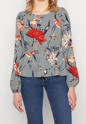 Femme portant un chemisier gris à manches longues avec des motifs floraux rouges et blancs, associé à un jean bleu, debout avec les bras détendus.