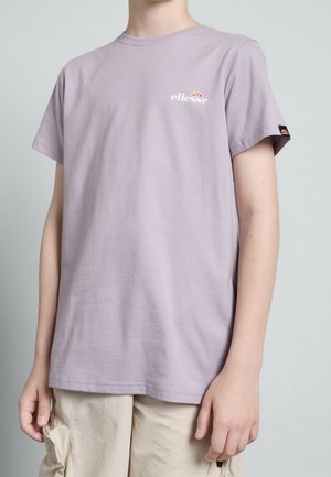 Giovane che indossa una t-shirt lavanda ellesse con logo sul petto e sulla manica, pantaloncini cargo beige, in piedi davanti a uno sfondo grigio uniforme.