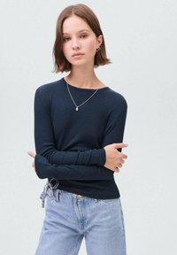 Námořnická modrá dlouhá rukávová halenka s žebrovanou texturou, vybavená kulatým výstřihem a rozšířenými manžetami, spárovaná s džínami ve světlém odstínu v pase na šňůrku.