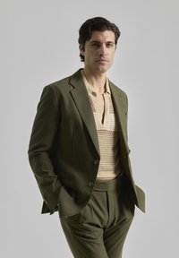Traje verde oliva a medida con chaqueta ajustada y pantalones a juego. Debajo, un top de punto beige que presenta un patrón geométrico en marrón y verde.