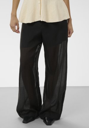 Personne portant un pantalon large noir transparent par-dessus un short noir et des chaussures noires pointues, associé à un haut crème boutonné.