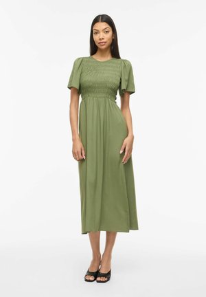 Femme aux longs cheveux noirs portant une robe midi vert olive avec des manches courtes bouffantes et un corsage froncé, associée à des talons ouverts noirs.