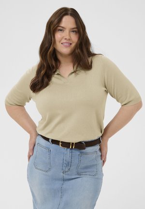Kvinde med langt brunt hår iført beige kortærmet top og lyseblå denimnederdel med sort bælte, smilende med hænderne på hofterne.