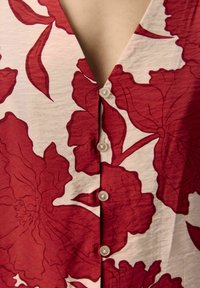 Prenda de color rojo y blanco con estampado floral que presenta un escote en V y cinco botones translúcidos en la parte frontal. Tejido texturizado con patrones visibles.