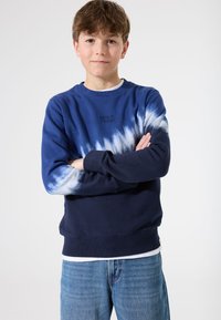 Sudadera azul y blanca con diseño de tie-dye, con puños y dobladillo acanalados. Presenta el texto "VOLVER EN 2 SEMANAS" en negro en la parte delantera. Combinada con jeans azules.