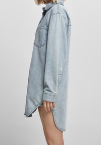 Vaaleansininen farkkumekko, jossa on teräväkauluksinen kaulus, pitkät hihat, yksi rintatasku ja epäsymmetrinen helma sivuhalkioilla.