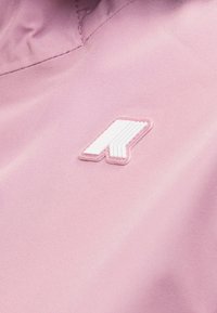 Κοντινή λήψη ροζ υφάσματος με ραμμένη λευκή και ροζ γεωμετρική λεπτομέρεια λογότυπου σε λεία υφασμάτινη επιφάνεια.